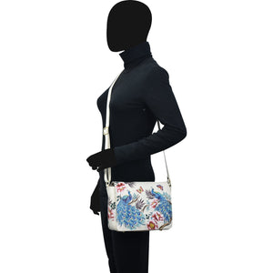 A person standing sideways wearing a black outfit and holding a hand painted Anuschka Flap Crossbody - 683 with a leather crossbody strap.
