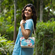 Load image into Gallery viewer, Woman smiling over shoulder with a hand-painted, floral-patterned Flap Crossbody - 683 by Anuschka featuring a leather crossbody strap in a lush garden.
