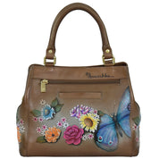 Close-up of a brown genuine leather handbag with hand-painted colorful flowers and a blue butterfly. The Multi Compartment Satchel - 690 features an adjustable shoulder strap, a gold zipper, and the brand name "Anuschka" visible near the top.