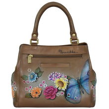 Load image into Gallery viewer, Close-up of a brown genuine leather handbag with hand-painted colorful flowers and a blue butterfly. The Multi Compartment Satchel - 690 features an adjustable shoulder strap, a gold zipper, and the brand name &quot;Anuschka&quot; visible near the top.
