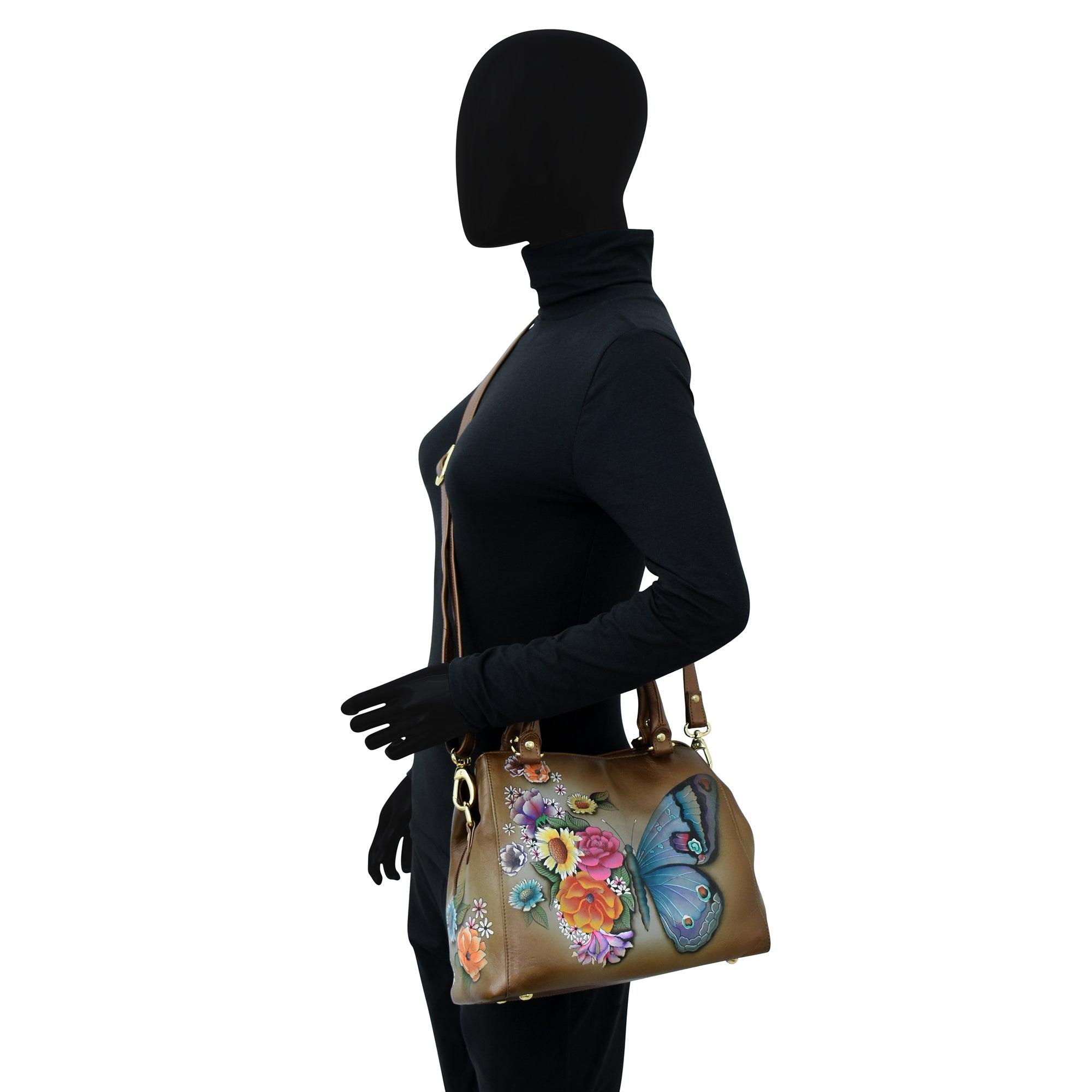 Silhouette of a woman dressed in black carrying the Anuschka Multi Compartment Satchel - 690, a brown handbag adorned with colorful butterfly and floral designs, featuring an adjustable shoulder strap.