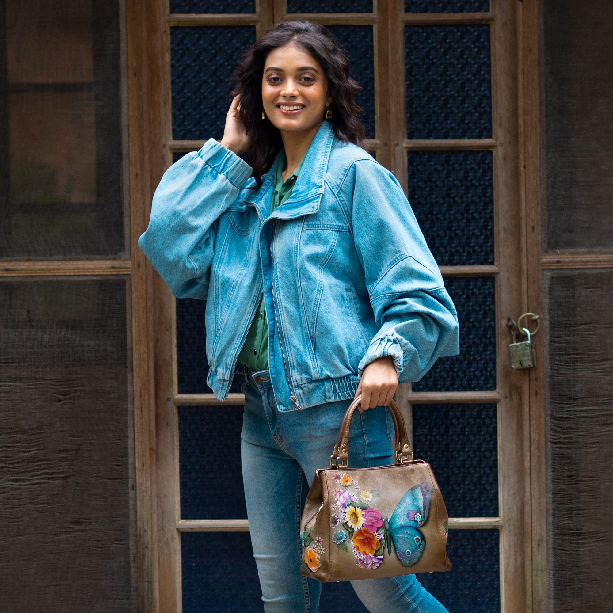 A person stands outside in front of a door, wearing a light blue denim jacket and jeans, and holding an Anuschka Multi Compartment Satchel - 690 with colorful designs. They are smiling and touching their hair.