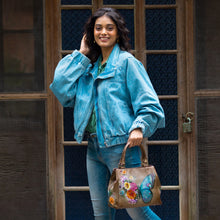 Load image into Gallery viewer, A person stands outside in front of a door, wearing a light blue denim jacket and jeans, and holding an Anuschka Multi Compartment Satchel - 690 with colorful designs. They are smiling and touching their hair.
