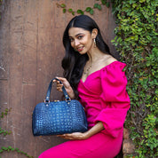 A woman in a pink off-the-shoulder dress holding an Anuschka Wide Organizer Satchel - 695 while posing against a wooden backdrop with green foliage.