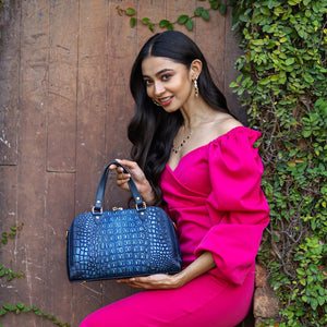 A woman in a pink off-the-shoulder dress holding an Anuschka Wide Organizer Satchel - 695 while posing against a wooden backdrop with green foliage.
