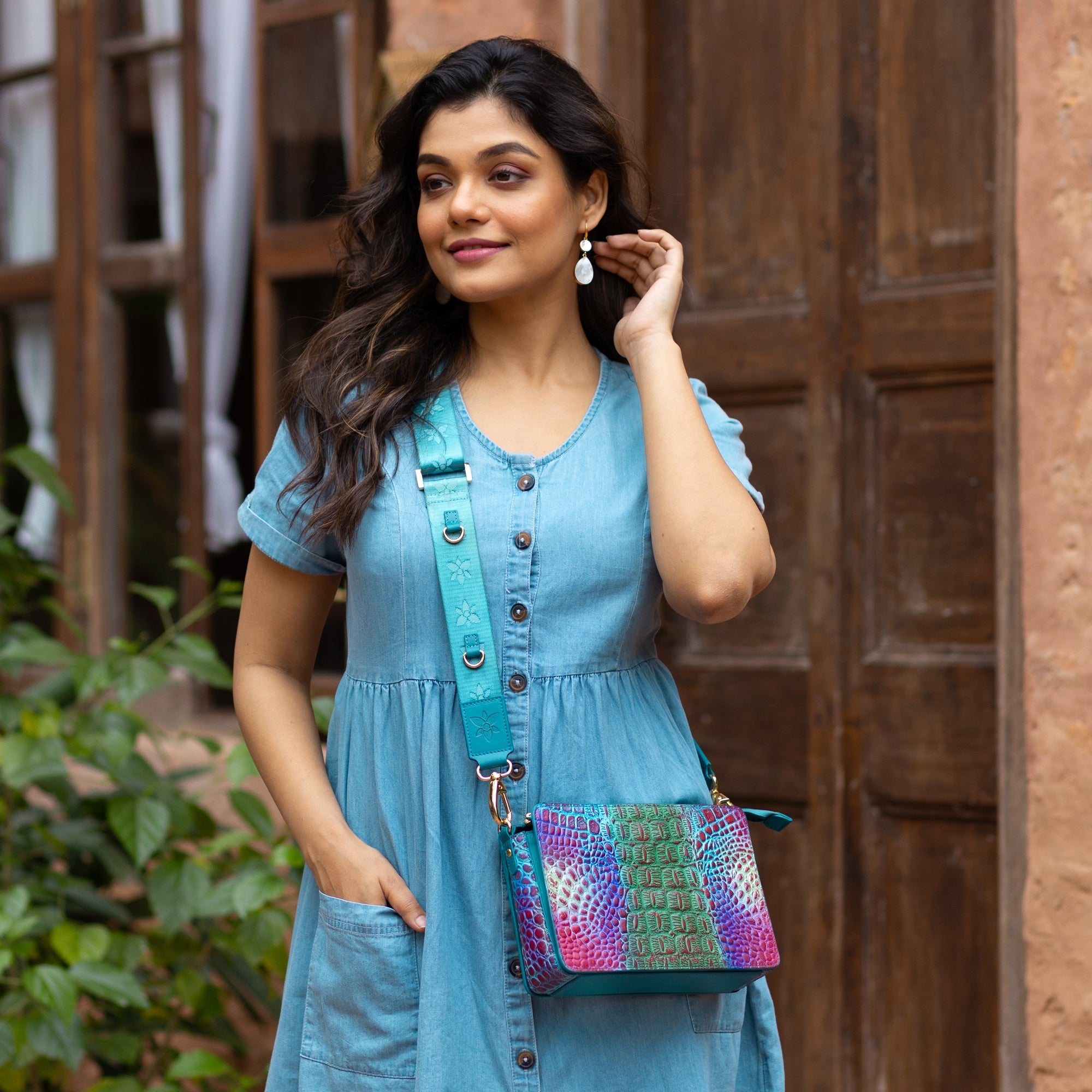 Woman in a blue dress with a colorful, organized Anuschka Triple Compartment Crossbody - 696 bag standing by a wooden door.