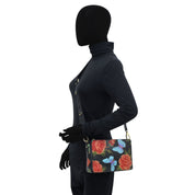 A mannequin dressed in black is showcasing the Anuschka Triple Compartment Crossbody - 696, a genuine leather bag adorned with a floral and butterfly pattern on a white background. The bag also features an adjustable strap for added convenience.