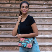 A woman wearing a black shirt and blue jeans stands on steps, holding an Anuschka Triple Compartment Crossbody - 696 with a floral pattern and an adjustable strap.