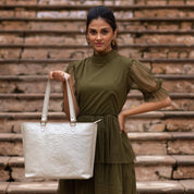 Woman posing with a Anuschka white embossed leather tote, wearing a green dress against a brick stair backdrop.