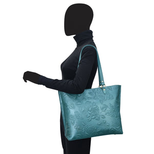 A mannequin with a black head and body wearing a dark long-sleeved top and carrying a large Anuschka Zip Top Tote - 698 with a floral pattern.