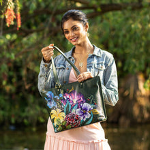 Load image into Gallery viewer, Woman smiling while showcasing an Anuschka Large Zip Top Tote - 698 with a floral-patterned, hand-painted artwork outdoors.
