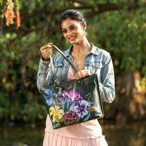 Woman smiling while showcasing an Anuschka Large Zip Top Tote - 698 with a floral-patterned, hand-painted artwork outdoors.