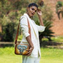 Load image into Gallery viewer, A woman in a white outfit poses outdoors with a detailed Anuschka Twin Top Messenger - 704 featuring an adjustable strap designed like an elephant.
