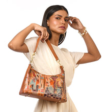 Load image into Gallery viewer, A woman in a white dress holds the Anuschka Zip-Top Shoulder Hobo - 707, which showcases a sophisticated leather design featuring an Egyptian motif and a chain detail shoulder strap.
