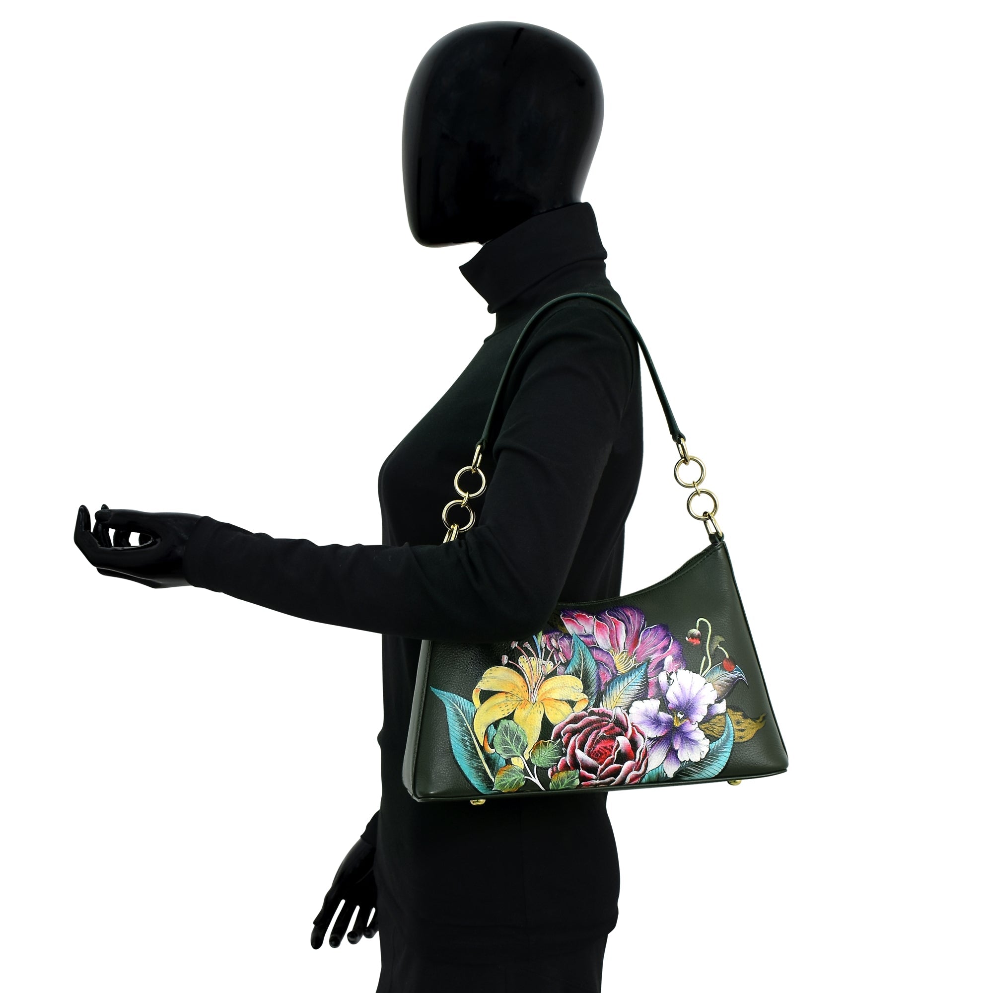 A mannequin in a black turtleneck and gloves holds an Anuschka Zip-Top Shoulder Hobo - 707, a dark green leather handbag adorned with colorful floral designs featuring pink, yellow, and purple flowers. This stylish handbag also boasts a chic chain detail shoulder strap.