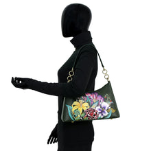 Load image into Gallery viewer, A mannequin in a black turtleneck and gloves holds an Anuschka Zip-Top Shoulder Hobo - 707, a dark green leather handbag adorned with colorful floral designs featuring pink, yellow, and purple flowers. This stylish handbag also boasts a chic chain detail shoulder strap.
