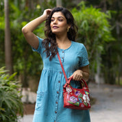 Woman in a blue dress carrying a floral red Anuschka satchel with crossbody strap outdoors.