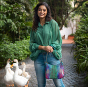 A woman smiling in a green blouse and blue jeans with an Anuschka Zip-Top Shoulder Hobo - 709 featuring chain link detail, standing in a garden pathway followed by a group of ducks.