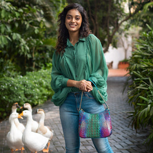 A woman smiling in a green blouse and blue jeans with an Anuschka Zip-Top Shoulder Hobo - 709 featuring chain link detail, standing in a garden pathway followed by a group of ducks.
