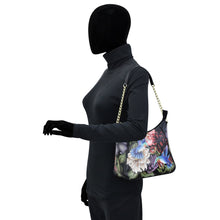 Load image into Gallery viewer, A mannequin wearing a black outfit and showcasing a Anuschka floral print leather zip-top shoulder hobo - 709 with a chain link shoulder strap.
