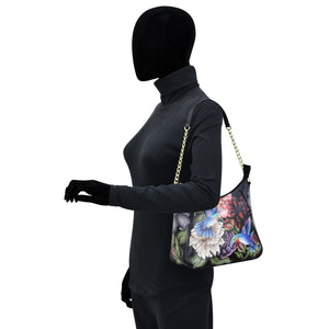 A mannequin wearing a black outfit and showcasing a Anuschka floral print leather zip-top shoulder hobo - 709 with a chain link shoulder strap.