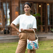 A woman standing outside a building with a hand on her hip, carrying an Anuschka Zip-Top Shoulder Hobo - 709 with a chain strap.