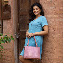 Load image into Gallery viewer, A smiling woman holding a pink Medium Everyday Tote - 710 with a leather exterior and zip entry stands beside a wooden door with a rustic wall background by Anuschka.
