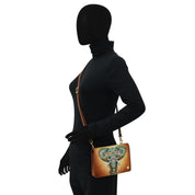 A person dressed in black from head to toe holds the Anuschka 4 in 1 Organizer Crossbody - 711, featuring a vibrant elephant design. The handbag is worn over the shoulder with a brown, genuine leather strap that can also be removed for crossbody wear.