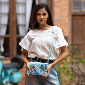 A woman standing in front of a rustic background, holding an Anuschka 4 in 1 Organizer Crossbody - 711 with a light blue strap.