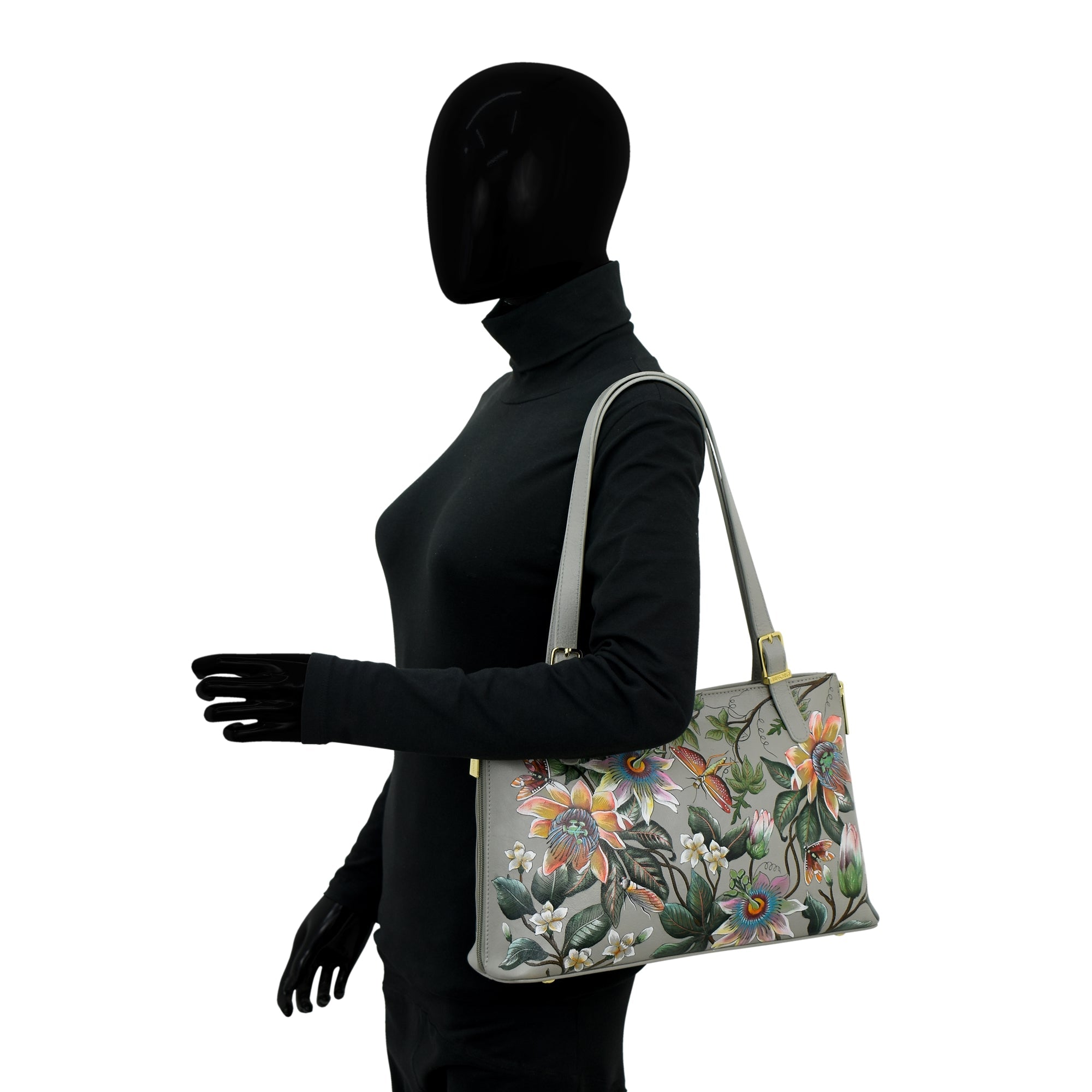 A mannequin in a black outfit holds the Anuschka Expandable Shopper Tote - 712, made of genuine leather and adorned with a colorful floral pattern.