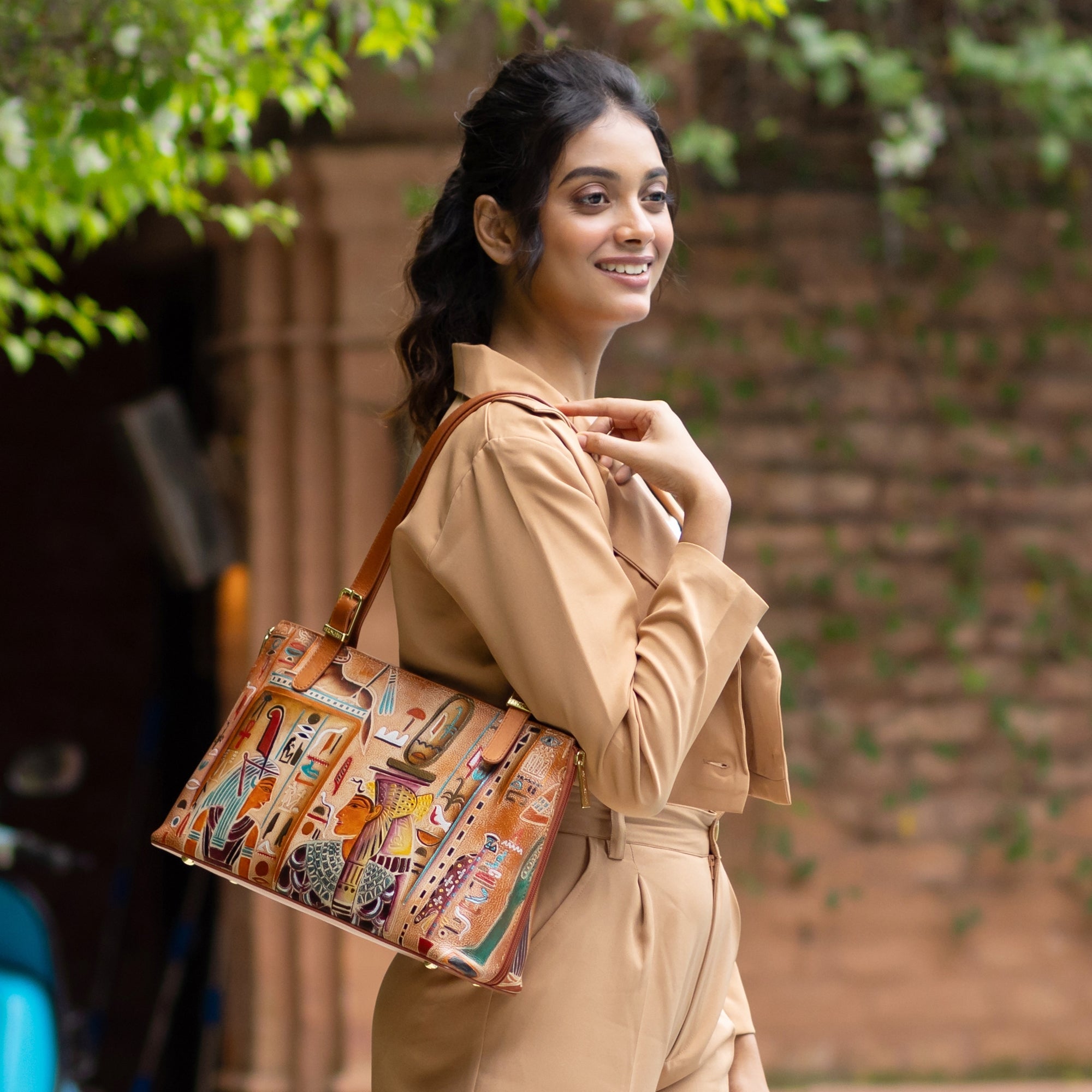 A woman smiles while posing outdoors, wearing a tan outfit and carrying the spacious Anuschka Expandable Shopper Tote - 712 adorned with colorful, intricate designs.