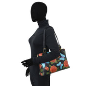 A mannequin wearing a black turtleneck holds an Anuschka Expandable Shopper Tote - 712, adorned with hand-painted artwork featuring red roses and blue butterflies.