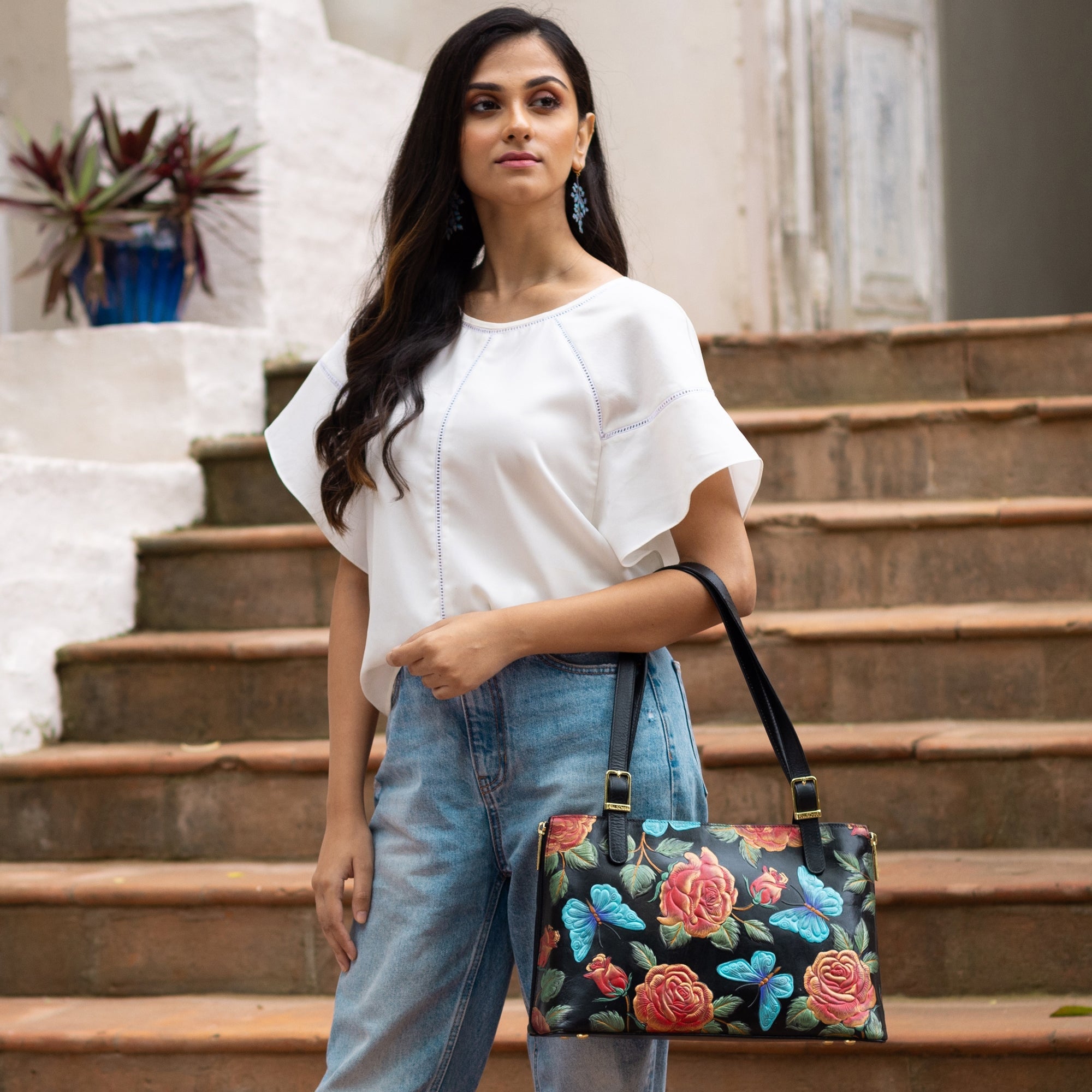 A woman standing on stairs wearing a white top, blue jeans, and holding an Anuschka Expandable Shopper Tote - 712 adorned with black floral patterns featuring red and blue flowers.