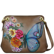 The Anuschka Crossbody Sling Bag - 713 is a brown genuine leather handbag showcasing a hand-painted design of vibrant flowers and a blue butterfly, complete with gold grommets and an adjustable crossbody strap.