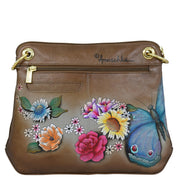 Anuschka’s Crossbody Sling Bag - 713 is a brown leather handbag adorned with colorful hand-painted artwork of flowers and a blue butterfly, featuring a zippered pocket, gold-tone hardware, and an adjustable crossbody strap.