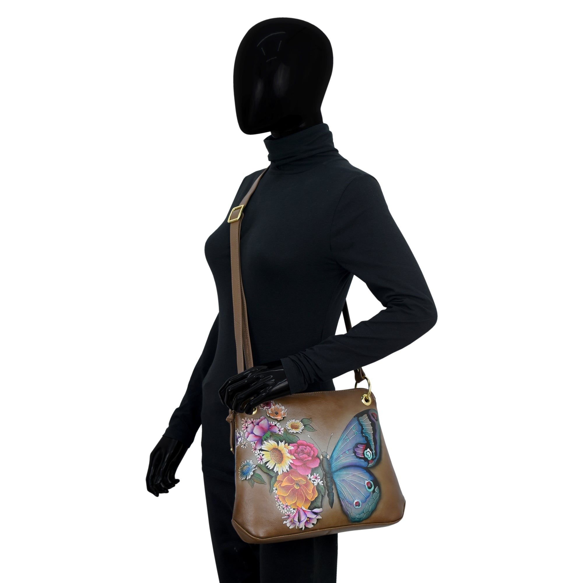 A mannequin in a black turtleneck and gloves showcases the Anuschka Crossbody Sling Bag - 713, crafted from genuine leather with hand-painted artwork featuring butterflies and floral designs, worn across the shoulder using its adjustable crossbody strap.