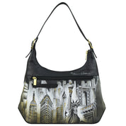 The Anuschka Slim Shoulder Hobo - 714 is a genuine leather handbag featuring a detailed cityscape design with the Statue of Liberty and various skyscrapers. It comes with a shoulder strap, front zipper pocket, gold-tone hardware accents, and an adjustable buckle handle for added convenience.