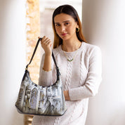 A woman wearing a light sweater holds the Anuschka Slim Shoulder Hobo - 714, featuring a cityscape design and made of genuine leather, standing in front of white columns.