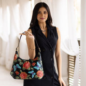 A woman stands indoors, holding the Anuschka Slim Shoulder Hobo - 714, a genuine leather handbag adorned with floral and butterfly designs. She wears a sleeveless black dress and jewelry, and has long dark hair. The modern Hobo bag features an adjustable strap, blending style with functionality.