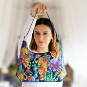 A person holds up the Slim Shoulder Hobo - 714 from Anuschka, an everyday handbag made of genuine leather, decorated with colorful illustrations of leopards and flowers. The focus is on the bag's modern Hobo design.