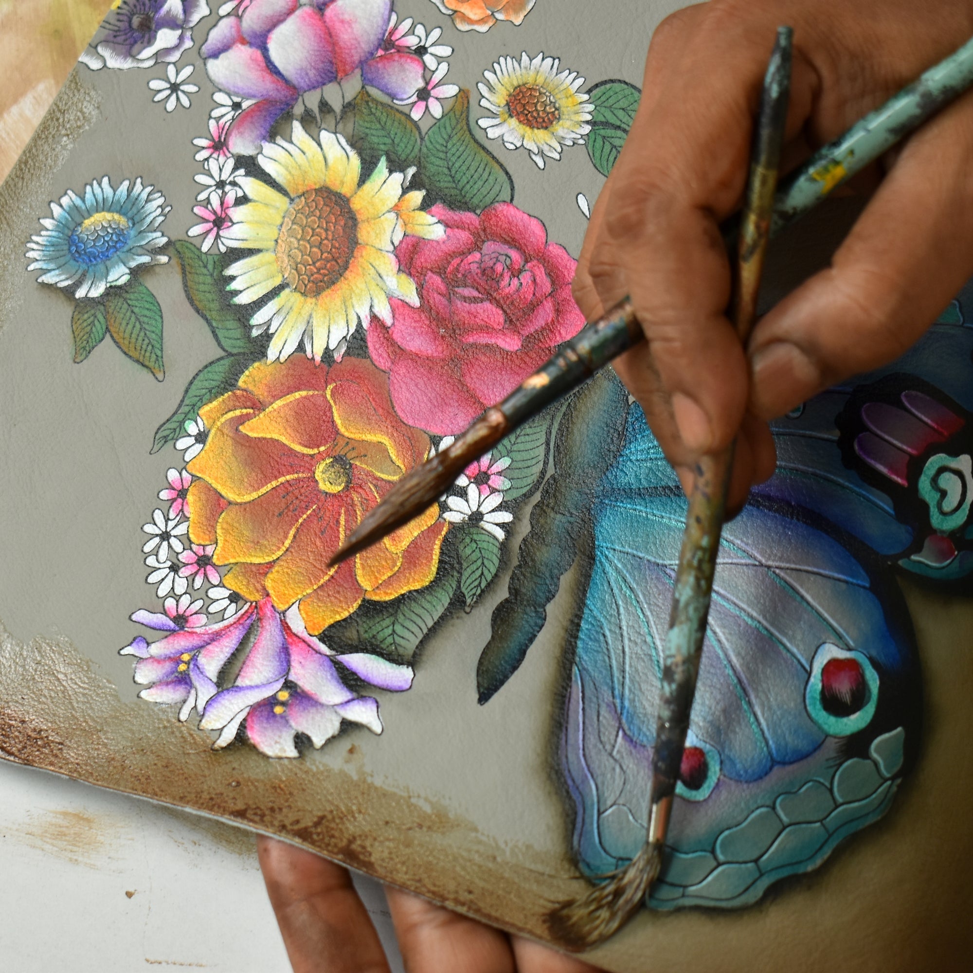 Close-up of hands painting a colorful image of flowers and a butterfly on a canvas, with Anuschka’s Multi Purpose Zip Pouch - 1182 in the background, holding minimalist essential art tools.