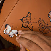 A person hand-painting butterflies on a Anuschka Classic Hobo With Side Pockets - 382 handbag with a shoulder strap.