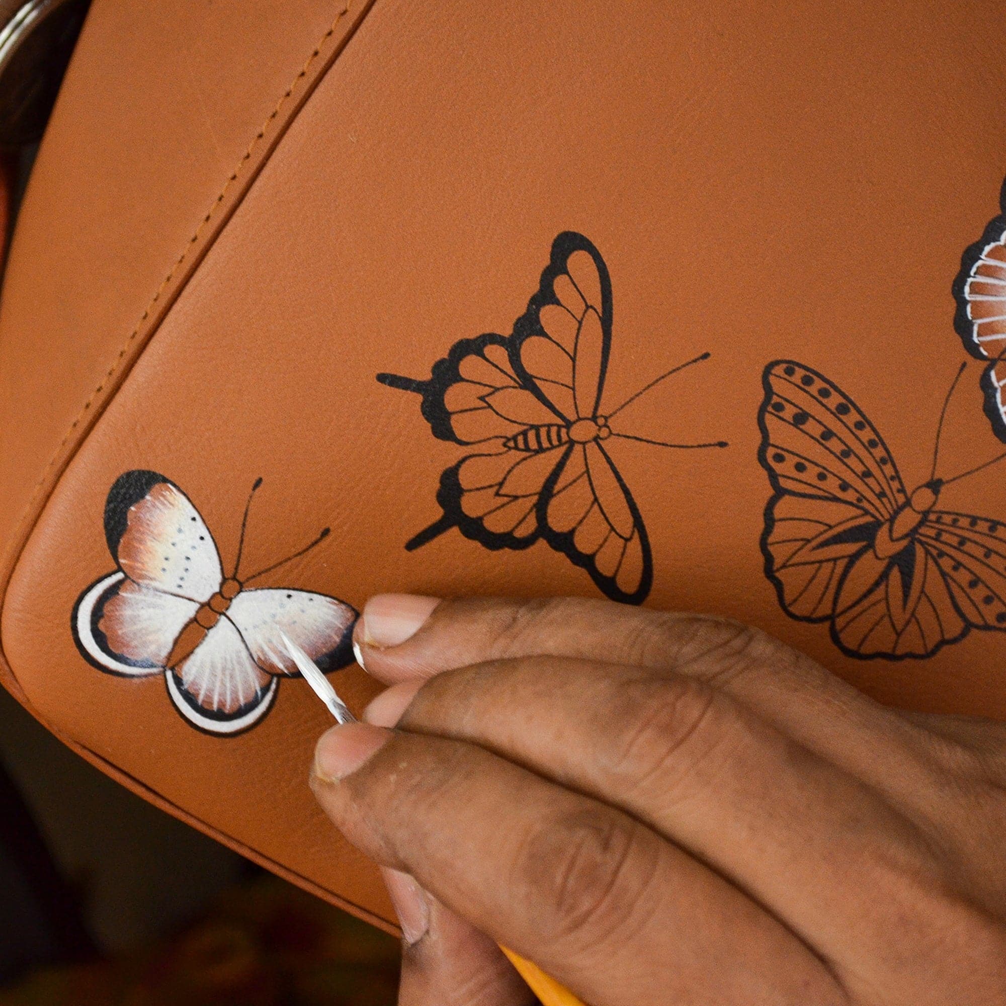 A person hand-painting butterflies on a Anuschka Classic Hobo With Side Pockets - 382 handbag with a shoulder strap.