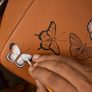 A person hand-painting butterflies on a Anuschka Classic Hobo With Side Pockets - 382 handbag with a shoulder strap.