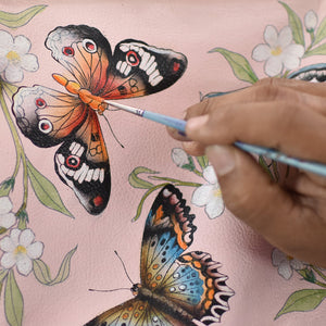 An artist's hand painting a detailed butterfly on a pink floral background, captured in hand painted artwork on an Anuschka Medium Zip-Around Eyeglass/Cosmetic Pouch - 1163.