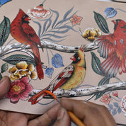 Close-up of a person creating a hand painted artwork on an Anuschka Card Organizer Wallet - 1184. The detailed image features red and yellow birds perched on branches, surrounded by various flowers and leaves, transforming the wallet into a unique credit card holder.