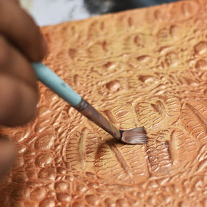 Person applying hand-painted designs to a textured leather Anuschka Large Zip Top Tote - 698 with a brush.