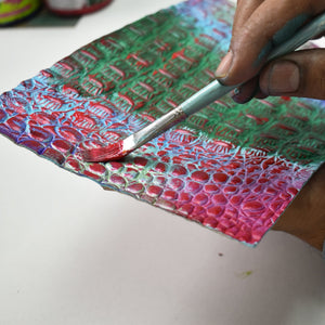Applying red paint to a textured Anuschka Three Fold Wallet - 1150 with a brush.