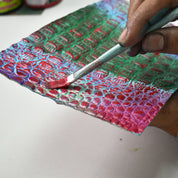 A person paints a textured surface with a paintbrush, applying red paint over a green and pink pattern reminiscent of an Anuschka Painted Leather Cuff - 1176.