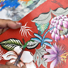 Load image into Gallery viewer, A person&#39;s hand painting a floral design on a textured red surface of an Anuschka Medium Zip-Around Eyeglass/Cosmetic Pouch - 1163.
